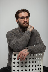 Wall Mural - Stylish handsome young hipster man with glasses with hairstyle and beard in vintage stylish knitted sweater sitting on white chairs in studio