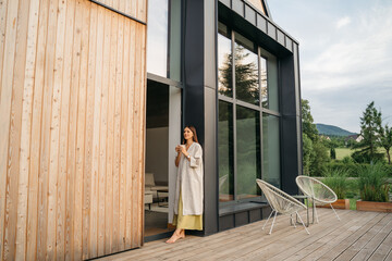 Young woman staying on terrace of wooden modern house with panoramic windows near pine forest. Concept of solitude and recreation on nature. Wellness and mindful resort. Beautiful place for vacation.