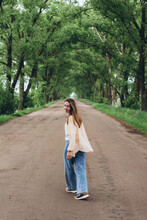 A Stylish Girl, A Woman Walks Through A Tunnel Of Green Trees. Warm Summer Day. The Girl, Lightly Dressed, Walks Off Into The Distance. Clothes Flutter In The Wind