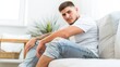 © Artsaba Family - A young man in torn jeans sits on a couch with a contemplative look, possibly reflecting on personal matters or simply taking a moment to rest. The light-filled room enhances the serene atmosphere.