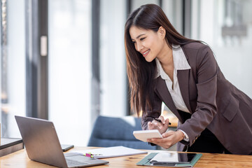 Image of young Asian beautiful joyful woman smiling while working with laptop in office do math finance on wooden desk, tax, accounting, statistics and analytical research concept