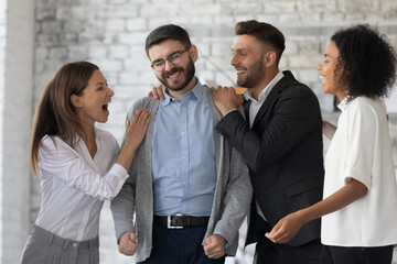 Congrats. Excited, smiling office workers congratulating their colleague after learning good news about his promotion. Cheering company employees hugging successful coworker in modern workspace