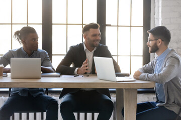 Wall Mural - Young businessmen discussing work issues sitting at desk with laptops in loft workspace. Entrepreneurs negotiating business deal. Friendly employees in modern office welcoming new coworker to the team