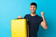 Young Colombian man isolated on blue background in vacation with travel suitcase