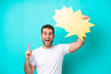 Wall Mural - Young caucasian man isolated on blue background holding an empty speech bubble with surprised expression