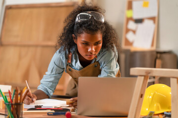 Young black African American woman carpenter concentrate on furniture design, sketching picture on paper blueprint while looking at laptop for reference. Craftsman working on table while drawing.