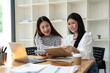 © Wasana - Two smiling female colleagues collaborate on paperwork with a laptop, notebooks in a contemporary office space.