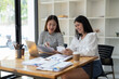 © Wasana - Two smiling female colleagues collaborate on paperwork with a laptop, notebooks in a contemporary office space.