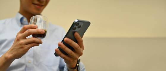 Poster - Cropped image of Young businessman holding mobile phone, typing text, chatting online, browsing the Internet while drinking a coffee, Banner background with copy space