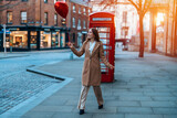 Fototapeta Londyn - young happy woman with a heart-shaped balloon falling a love, having a fun day, walking against the red phone box in English city Spring is in the air Lifestyle, tourism, valentines day concept