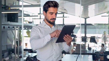 Wall Mural - Busy young Latin business man entrepreneur using tablet standing in office at work. Male professional executive manager using tab computer managing financial banking tech data.