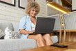 © puhhha - Cheerful curly-haired young woman using a laptop on a cozy sofa at home, embodying a relaxed work-from-home lifestyle.
