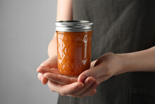 Woman Holding Glass Jar Of Delicious Persimmon Jam On Gray Background, Closeup. Space For Text