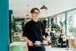 © okrasiuk - Portrait of young smiling affable waiter carrying on tray with coffee cups to client table in cafe. Hospitality service job. Occupation and work concept. Part-time work for students. Selective focus.