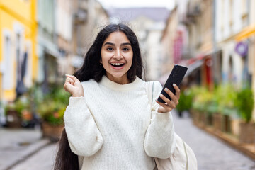 Portrait of a happy and excited young Indian girl in a white sweater standing in the middle of a city street and holding a phone in her hands, enjoying success and news on camera