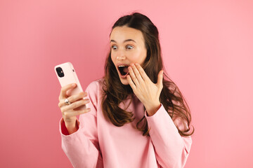 Shocked brunette woman wear pink long sleeves top hold mobile phone isolated on pink background studio. Shocked woman with open mouth, hand near face wow win emotions. Amazing useful app. OMG.