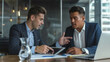 © MP Studio - Two professionals engaged in a serious discussion with one holding a tablet, sitting at a wooden table in a modern office environment, with a laptop and various documents laid out in front of them.