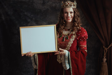 Wall Mural - smiling medieval queen in red dress showing blank board