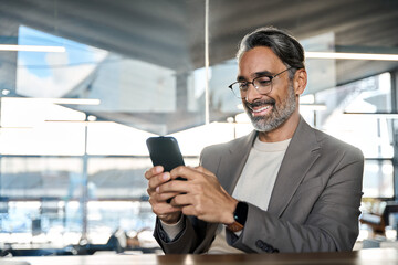 Wall Mural - Smiling mature business man executive sitting at desk using smartphone. Happy busy middle aged businessman entrepreneur investor looking at mobile cell phone working in office holding cellphone.