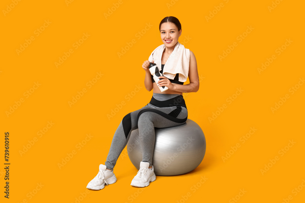 Young woman in sportswear and with bottle of water sitting on fitball against yellow background