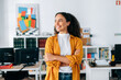 © Kateryna - Positive successful confident beautiful hispanic curly haired business woman in orange shirt, business coach, manager, standing in a modern office with arms crossed, looking away and smiling