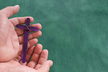 Wall Mural - Male hand holding purple wooden cross flat lay in green background. Holy week and lent season celebration concept.