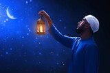 Fototapeta Sport - Young asian muslim man with beard holding arabic lantern looking at beautiful blue night sky with stars and crescent moon