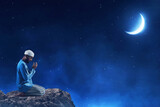 Fototapeta Sport - Young asian muslim man with beard praying , sitting on on top rock monuntain at beautiful blue night sky with stars and moon