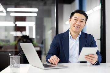 Wall Mural - Portrait of a smiling and successful young Asian man in a suit working in an office center, sitting at a desk, using gadgets and looking confidently at the camera.