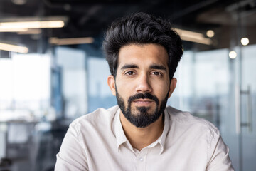 Wall Mural - Close-up photo of a serious young Indian man with a beard and a shirt working in the office, sitting and looking confidently into the camera