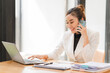 © Kritdanai - A portrait of a beautiful and confident Asian businesswoman in a pink business suit sits at her desk in the office with her arms crossed, smiling at the camera.