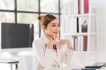 A portrait of a beautiful and confident Asian businesswoman in a pink business suit sits at her desk in the office with her arms crossed, smiling at the camera.