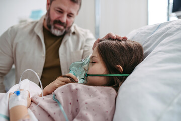 Father holding hand of his daughter in hospital bed. Child patient with an oxygen mask in ICU.