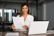 © Stock 4 You - Smiling European Latin business woman accountant holding documents, work at laptop pc doing online trade market analysis. Portrait 40s hispanic businesswoman with paperwork in office looking at camera