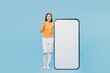 © ViDi Studio - Young woman of Asian ethnicity wear orange t-shirt casual clothes big huge blank screen mobile cell phone smartphone with workspace copy space point index finger up isolated on plain blue background.