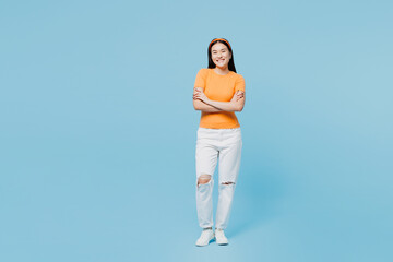 Wall Mural - Full body smiling happy young woman of Asian ethnicity she wear orange t-shirt casual clothes hold hands crossed folded isolated on pastel light blue cyan background studio portrait Lifestyle concept.