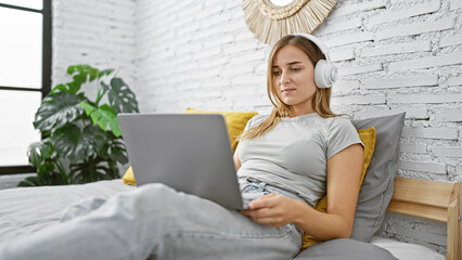 Poster - Young blonde woman using laptop and headphones sitting on bed at bedroom