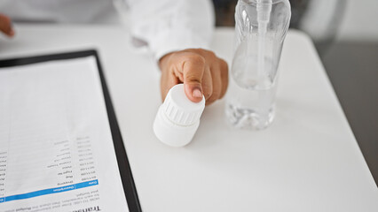 Healthcare professional in white lab coat opens medication bottle at clinic desk near water and digital tablet.