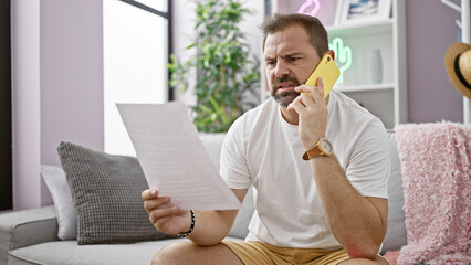 Wall Mural - Mature hispanic man with grey hair reading a document and talking on a yellow phone in a modern living room.