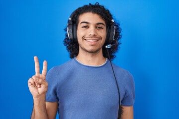 Poster - Hispanic man with curly hair listening to music using headphones showing and pointing up with fingers number two while smiling confident and happy.