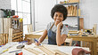 Smiling african american woman taking notes in a sunny carpentry workshop, surrounded by tools and timber.