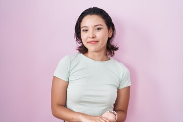 Wall Mural - Hispanic young woman standing over pink background with hands together and crossed fingers smiling relaxed and cheerful. success and optimistic