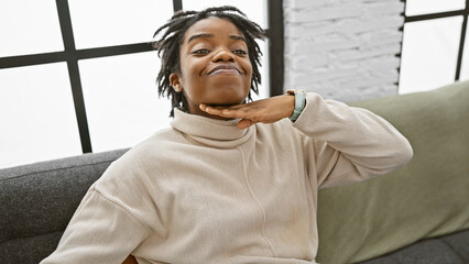 Poster - Portrait of a young woman with dreadlocks making a cutthroat gesture indoors on a sofa.