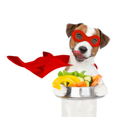 Poster - Licking lips Jack russell terrier puppy wearing superhero costume holds bowl of vegetables and looks at camera. Isolated on white background