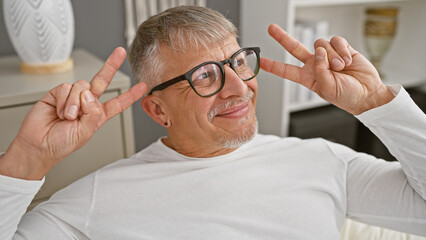 Sticker - Smiling middle-aged man with glasses making peace sign while relaxing in a white bedroom