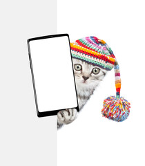 Aufkleber - Cute kitten wearing warm hat with pompon holds smartphone with white blank screen in it paw above empty white banner. isolated on white background