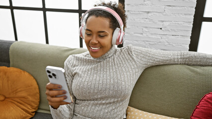 Poster - An adult woman enjoys music on headphones while holding a smartphone, sitting on a sofa in a cozy living room.