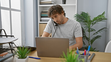 Poster - A focused man working and multitasking with a laptop and telephone in a modern office setting.