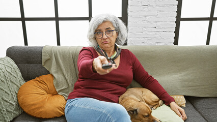 Poster - A mature woman with grey hair sits on a couch next to a dog, offering a remote control indoors.