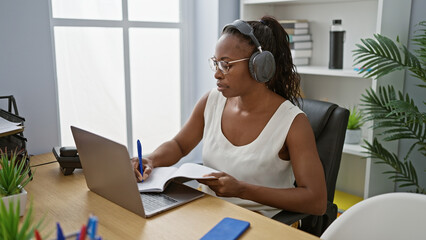 Sticker - African american woman wearing headphones taking notes at her office desk indoors